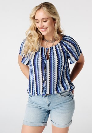 Femme souriante aux longs cheveux blonds porte un chemisier bleu à motifs zigzag et un short en jean clair, debout les mains derrière le dos.