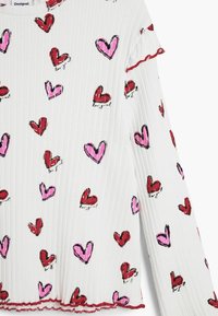 Biely rebrovaný dlhý rukáv s ružovo-červeným srdcovým vzorom, ozdobený červenou volánovou lemovkou na manžetách a výstrihu.