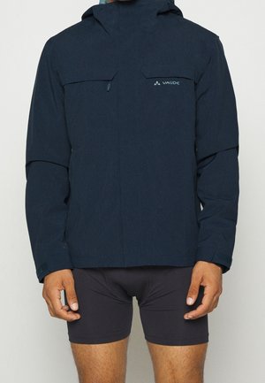 Mann trägt eine marineblaue Kapuzenjacke mit Vaude-Logo und schwarze Radsportshorts vor einfachem Hintergrund.