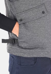 Giacca esterna grigia e strutturata con motivo a spina di pesce, dotata di bottoni a pressione neri e di una fascia regolabile. Abbinata a pantaloni blu navy.