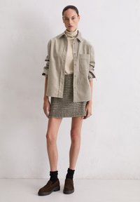 Lichtgrijs oversized overhemd, beige coltrui, pied-de-poule minirok, bruine instapschoenen, zwarte geribbelde sokken; effen achtergrond.