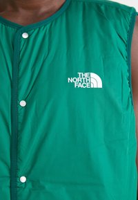 Chaqueta sin mangas verde con botones a presión, usada por una persona, que muestra el logo blanco "The North Face" en el pecho.