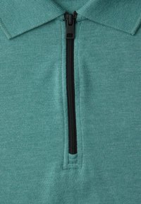 Zelená látková košile s tyrkysovým límečkem a černým zipem uprostřed částečně rozepnutým vpředu.