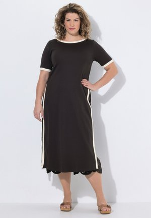 Kvinde med krøllet hår iført en sort midi-kjole med hvide kanter, stående med den ene hånd på hoften og iført lysebrune sandaler mod en ensfarvet baggrund.