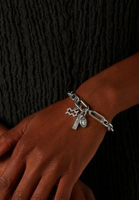 Zilveren charmearmband met grote schakels, decoratieve bedels en een gestructureerde ketting, gedragen op een gestructureerde donkere stoffen achtergrond.
