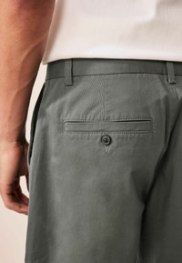 Graue Hose mit glatter Textur, ausgestattet mit einer Gesäßtasche mit einem einzelnen Knopf und Gürtelschlaufen, kombiniert mit einem hellen Hemd.