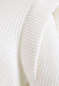 Vit stickad tröja med ribbad textur, bred krage och avslappnad passform. Närbild visar sömnadsdetaljer och mjukt, chunky garn.