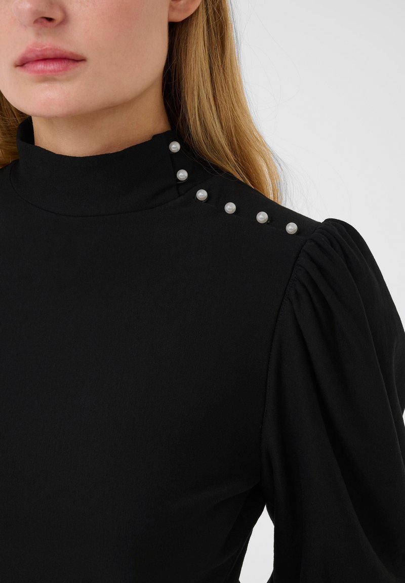 Camisa negra con cuello alto, mangas abullonadas y botones decorativos de perla en el hombro. La tela parece suave y ligeramente elástica.