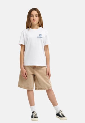 Biała bawełniana koszulka z małym niebieskim logo graficznym, zestawiona z beżowymi szortami dżinsowymi, białymi skarpetkami i czarnymi trampkami. Pozuje na stojąco.