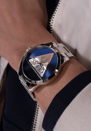 Montre en acier inoxydable argenté avec un cadran bleu, un accent triangulaire argenté et des aiguilles métalliques. Présentée sur un poignet, arborant une texture lisse.