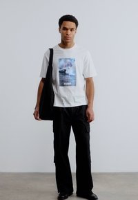 Νέος άνδρας που φοράει λευκό T-shirt με γραφικά, μαύρο παντελόνι cargo, μαύρα παπούτσια και κρατάει μια μεγάλη μαύρη τσάντα ώμου, στέκεται σε απλό φόντο.