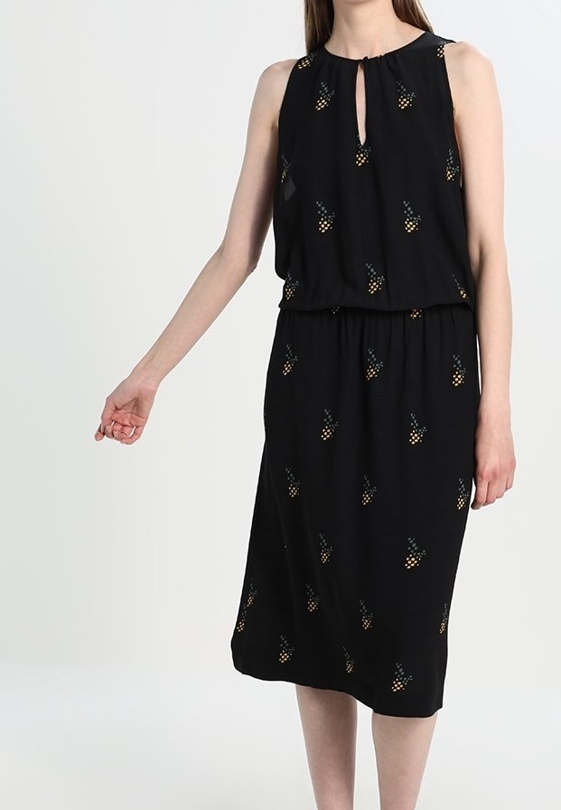 Femme portant une robe midi noire sans manches avec de petits motifs d’ananas jaunes et verts sur un fond uni clair.