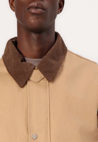 Beige jacket with a brown corduroy collar, featuring a button closure and a smooth texture. Visible stitching detail enhances design.