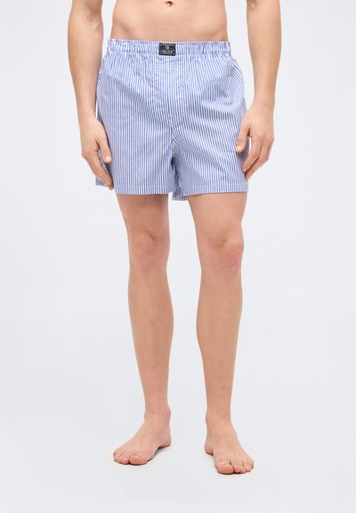 Άνδρας που φοράει μπλε και λευκά μποξεράκια με κάθετες ρίγες, στέκεται ξυπόλητος μπροστά σε έναν απλό φωτεινό φόντο.