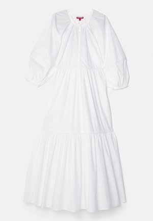 Frau trägt ein langes weißes Stufenkleid mit gerafften Ärmeln und Knöpfen, kombiniert mit roten Stiefeletten mit genietetem Profil.