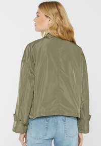 Olivgrüne leichte Jacke mit lässiger Passform, abgesenkten Schultern und hochrollbaren Ärmeln. Sichere Textur mit minimalen Details.