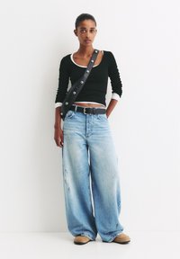 Tânără stând în picioare, purtând un top negru cu mâneci lungi, blugi largi, albaștri deschiși, încălțăminte maro deschis și o geantă neagră crossbody cu nituri argintii.