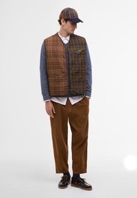 Hnědá kostkovaná péřová vesta s knoflíkovým zapínáním, nošená přes modrou košili s dlouhým rukávem a doplněná hnědými manšestrovými kalhotami a černými botami.
