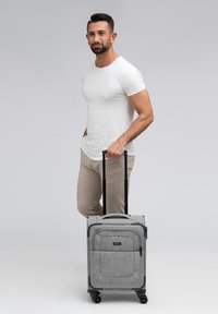Grijze wheeled carry-on koffer met een textuurstof buitenkant, meerdere zakken en een uitschuifbare handgreep, geplaatst naast een staande man.