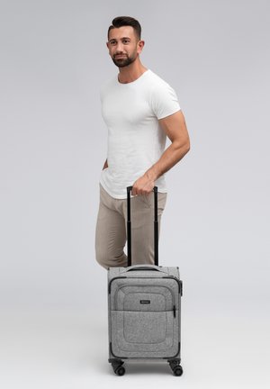 Grijze wheeled carry-on koffer met een textuurstof buitenkant, meerdere zakken en een uitschuifbare handgreep, geplaatst naast een staande man.
