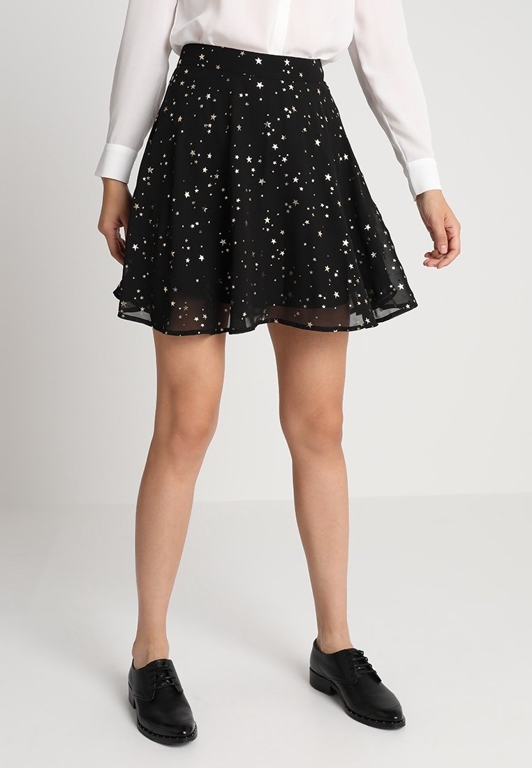 Femme portant une jupe noire avec des motifs d'étoiles blanches et dorées, un chemisier blanc manches longues et des chaussures noires à lacets, debout sur un fond uni.