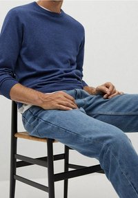 Jersey de punto azul marino con cuello redondo, combinado con jeans de mezclilla azul claro, sentado en una silla de madera negra con asiento trenzado.