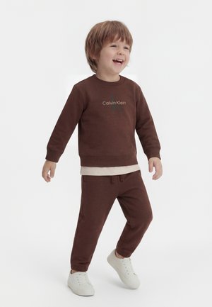 Jeune garçon souriant marchant, portant un sweat-shirt marron Calvin Klein, un pantalon assorti, une chemise blanche en dessous et des baskets blanches sur fond blanc.