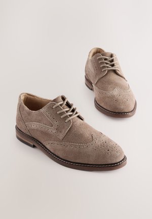 Scarpe stringate beige in suede con dettagli brogue, punta arrotondata e suola in pelle marrone. La parte superiore presenta perforazioni e cuciture a contrasto.