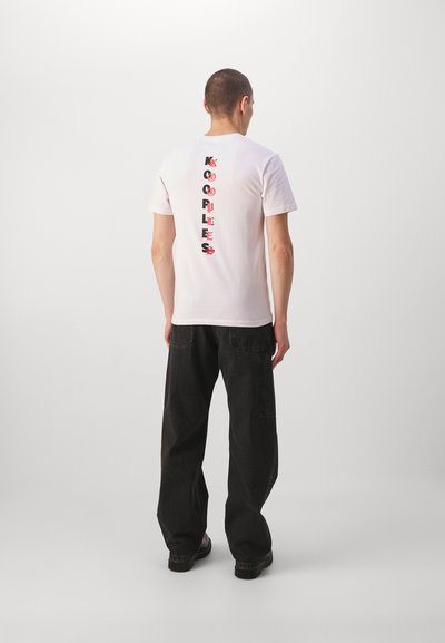 Camiseta blanca con texto vertical rojo y negro "KOOPLES" en la parte trasera. Lleva puesta con pantalones negros de corte holgado y calzado oscuro.