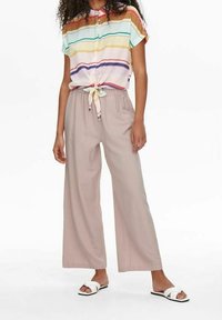 Kvinna som bär en kortärmad, knappad, randig skjorta knuten i midjan tillsammans med beige vida byxor och vita halksandaler.