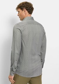 Mann, der von hinten in einem eng anliegenden grauen Langarmhemd mit Muster und olivgrüner Hose vor einem schlichten weißen Hintergrund zu sehen ist.