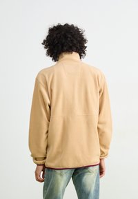 Beige fleece pullover with a high collar and long sleeves. Features a back yoke detail and a maroon hem, worn over light-wash jeans.