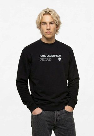 Mladý muž so svetlými vlasmi v čiernej mikine Karl Lagerfeld Jeans a tmavosivých džínsoch, stojaci s rukami v kapse.