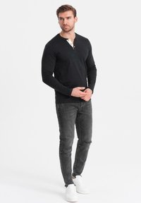 Maglietta henley nera a maniche lunghe con colletto abbottonato, abbinata a jeans grigi strappati e sneakers bianche. Design semplice, aderente e texture liscia.