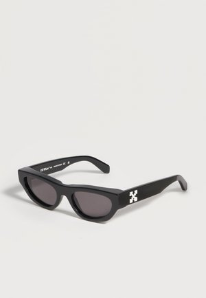 Lunettes de soleil rectangulaires noires avec des verres foncés et un détail de logo blanc sur les branches, reposant sur une surface blanche.