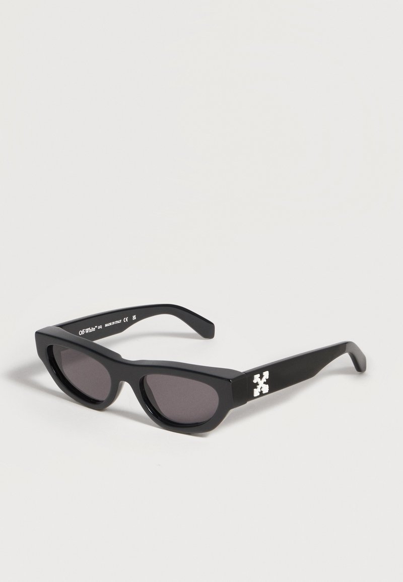 Lunettes de soleil rectangulaires noires avec des verres foncés et un détail de logo blanc sur les branches, reposant sur une surface blanche.