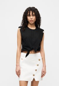 Μαύρο φανελάκι κομμένο, με δεμένο μπροστά και διακοσμήσεις στους ώμους· συνδυασμένο με λευκή ζώνη-φούστα με χρυσά κουμπιά και διαγώνιο τελείωμα.