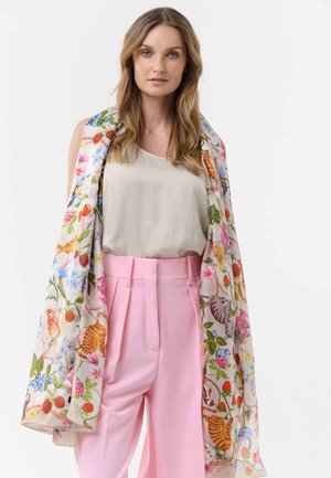 Woman with long hair wears a beige top, high-waisted pink pants, and a colorful floral and animal patterned scarf draped over her shoulders.