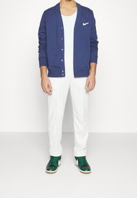 Marinblå cardigan med knappar och en liten turkos Nike-logotyp, i kombination med vita byxor och gröna sneakers med vita detaljer.