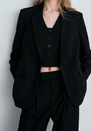 Frau trägt einen schwarzen oversized Blazer, geknöpfte schwarze Cropped-Weste und schwarze hoch taillierte Hose vor schlichtem Hintergrund.