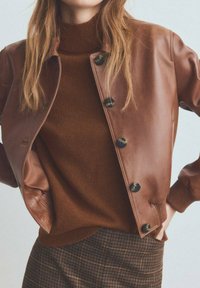 Veste aviateur en cuir marron avec fermeture à boutons, col et poignets côtelés. En dessous, un pull marron associé à un tissu de jupe à carreaux.
