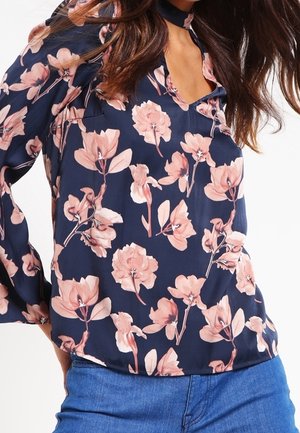 Vrouw draagt een marineblauwe blouse met roze bloemenprint en een keyhole-halslijn, gecombineerd met blauwe spijkerbroek.
