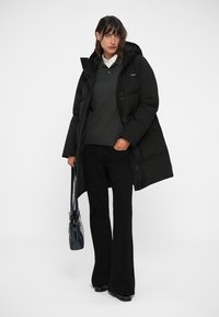 Casaco puffer preto com capuz, apresentando um fecho de correr na frente e bolsos com aba, combinado com um suéter cinza e calças pretas largas, segurando uma bolsa preta.