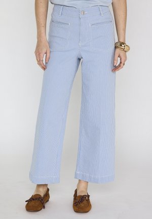 Vrouw die lichtblauwe en witte wijde broek met verticale strepen en voorzakken draagt, gecombineerd met bruine mocassins en gouden sieraden.