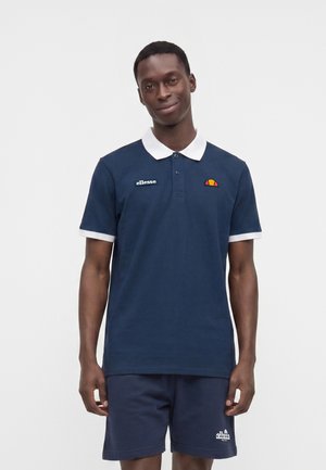 Homme portant un polo ellesse bleu marine avec col blanc et un short bleu marine assorti, debout devant un fond blanc uni.