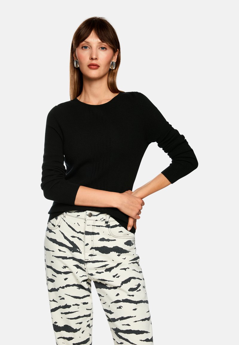 Black ribbed sweater with long sleeves, paired with white jeans featuring a black abstract pattern. Model has styled hair and large earrings.