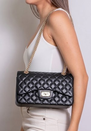 Donna con canotta bianca e pantaloni beige che porta una borsa in pelle nera trapuntata con tracolla a catena dorata sulla spalla.