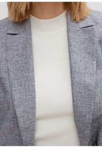 Persona che indossa un blazer grigio dalla texture particolare sopra un top bianco a coste con collo alto, accorciato al collo e al petto.