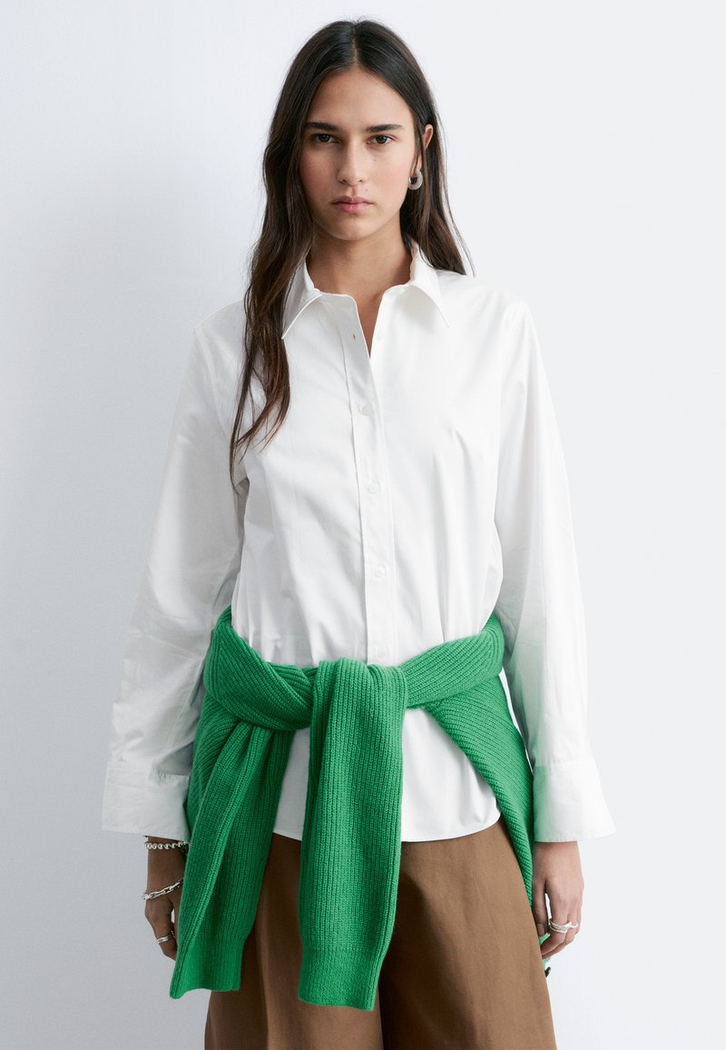 Young woman wearing a white button-up shirt, green sweater tied around waist, and brown pants, standing against a plain light background.