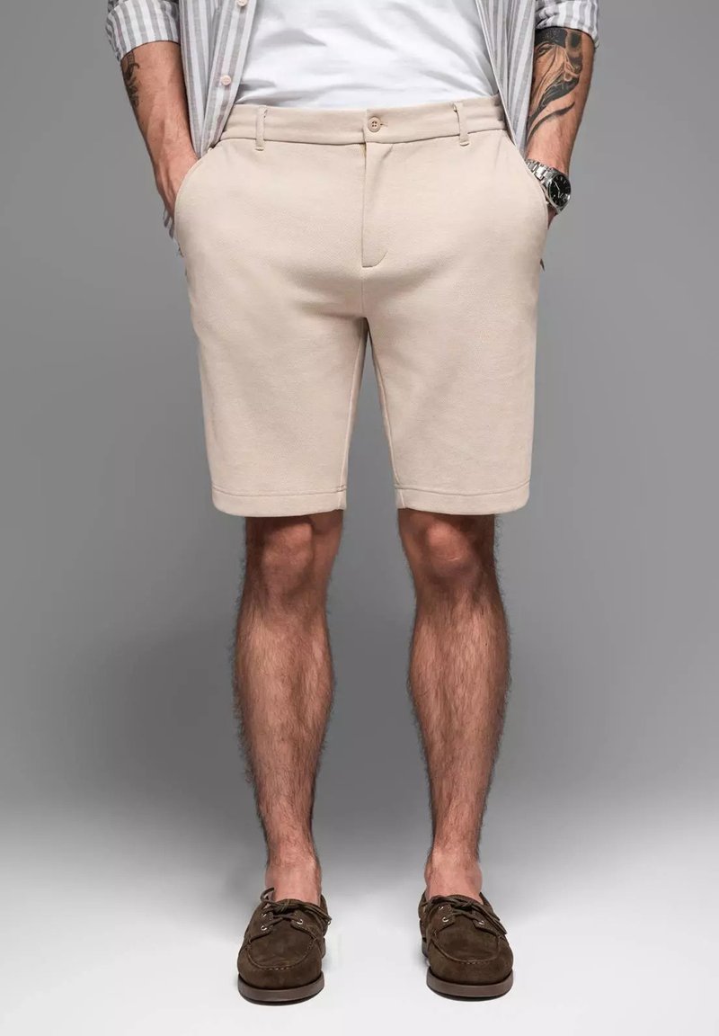 Pantalones cortos beige hechos de una tela texturizada, con un cierre de botón, bolsillos laterales y un corte recto, combinados con zapatos de ante marrones.
